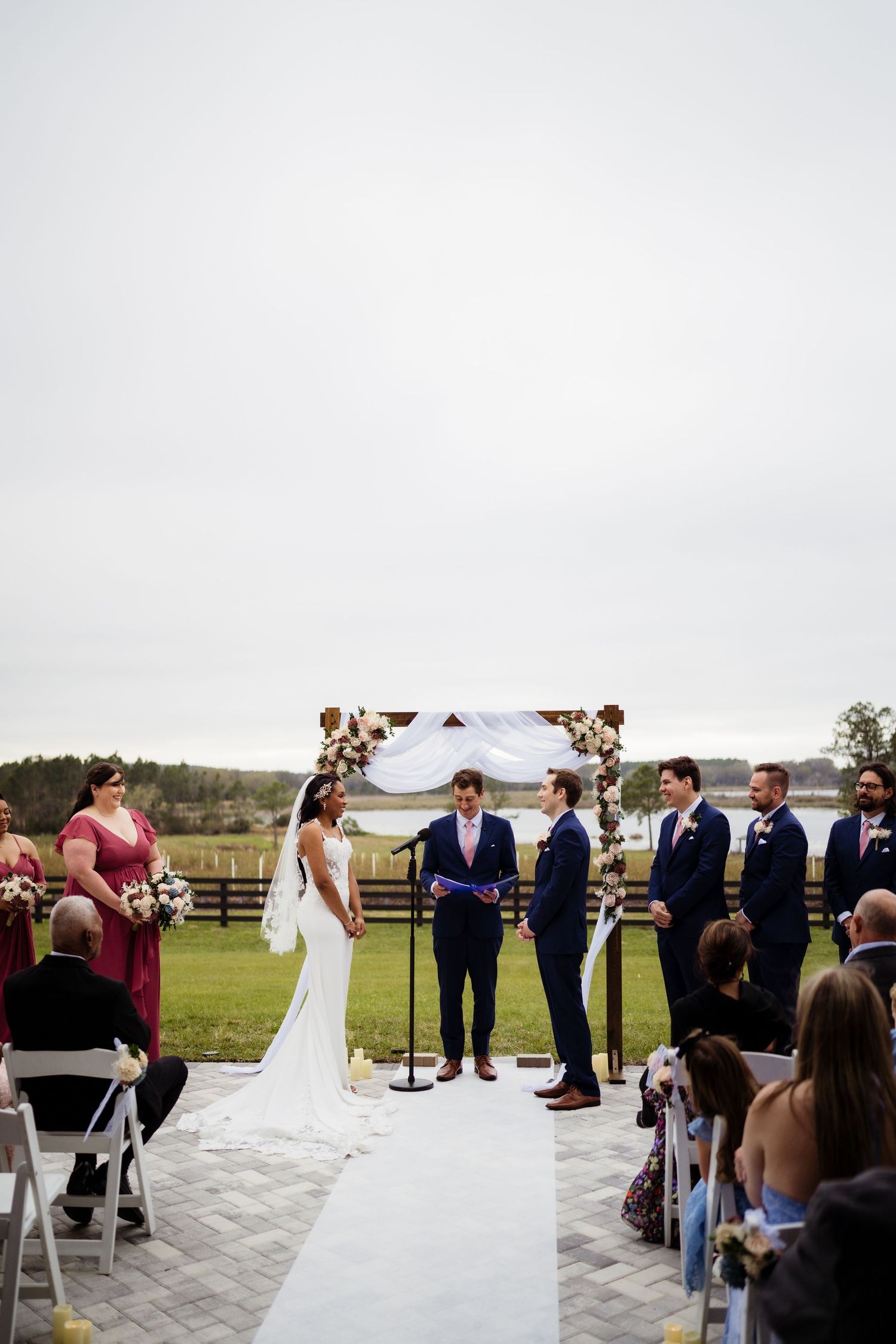 An outdoor wedding ceremony unfolds on a stone patio with guests seated in white chairs and rolling hills in the background.