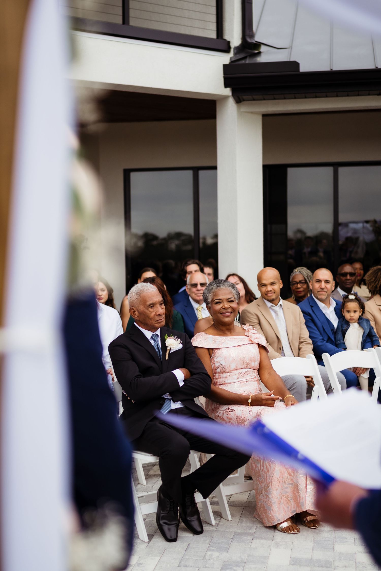 Wedding guests seated outdoors in white chairs at a courtyard ceremony venue with white columns in background.
