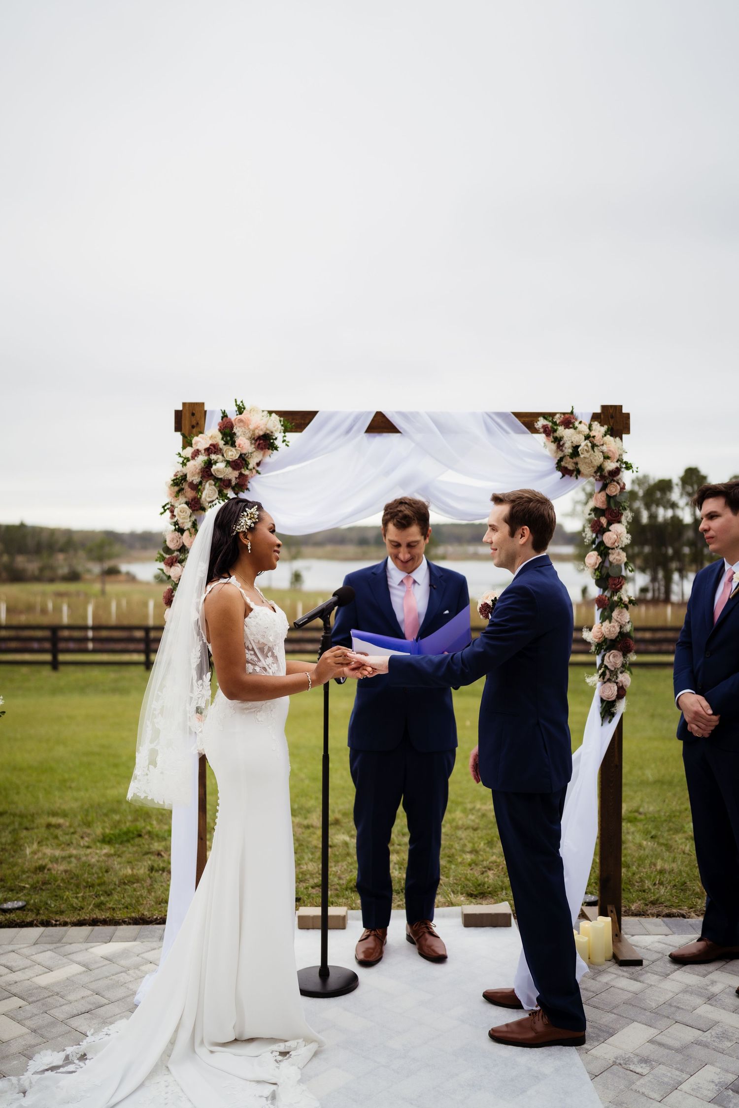 Wedding ceremony taking place outdoors on stone patio with white arch and mountain views in background.