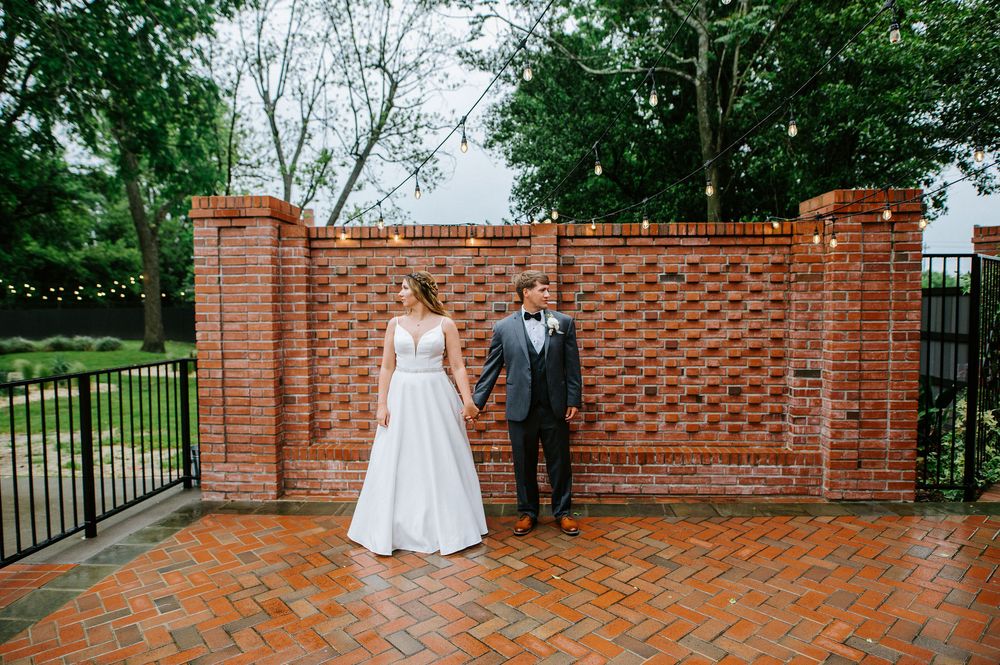 A couple stands together on a brick patio in front of a decorative woven brick wall with string lights overhead.