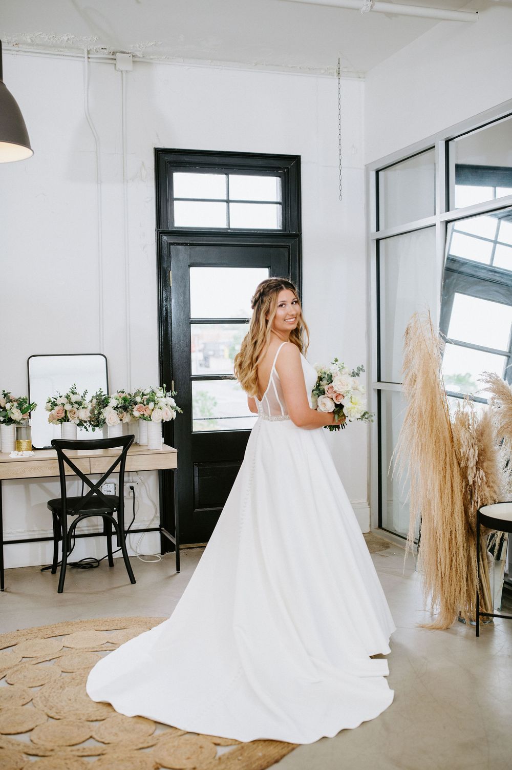 A bride in a flowing white wedding gown twirls gracefully in a bright, minimalist studio space with dark-framed windows.