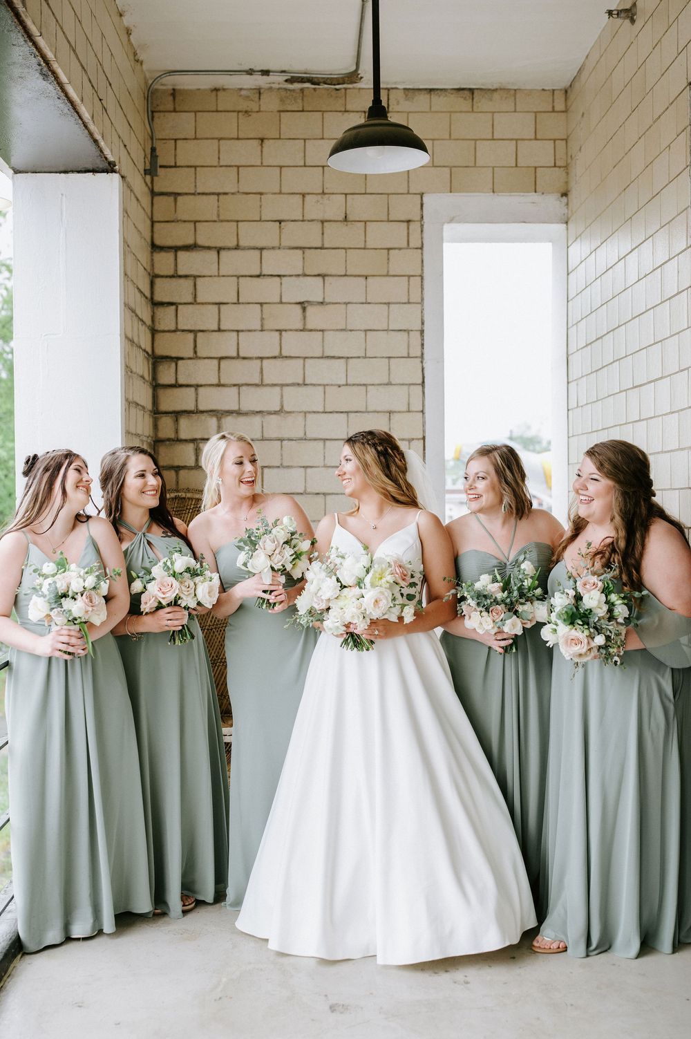 Bridesmaids in sage green dresses hold white bouquets while standing with bride in white gown against brick wall.