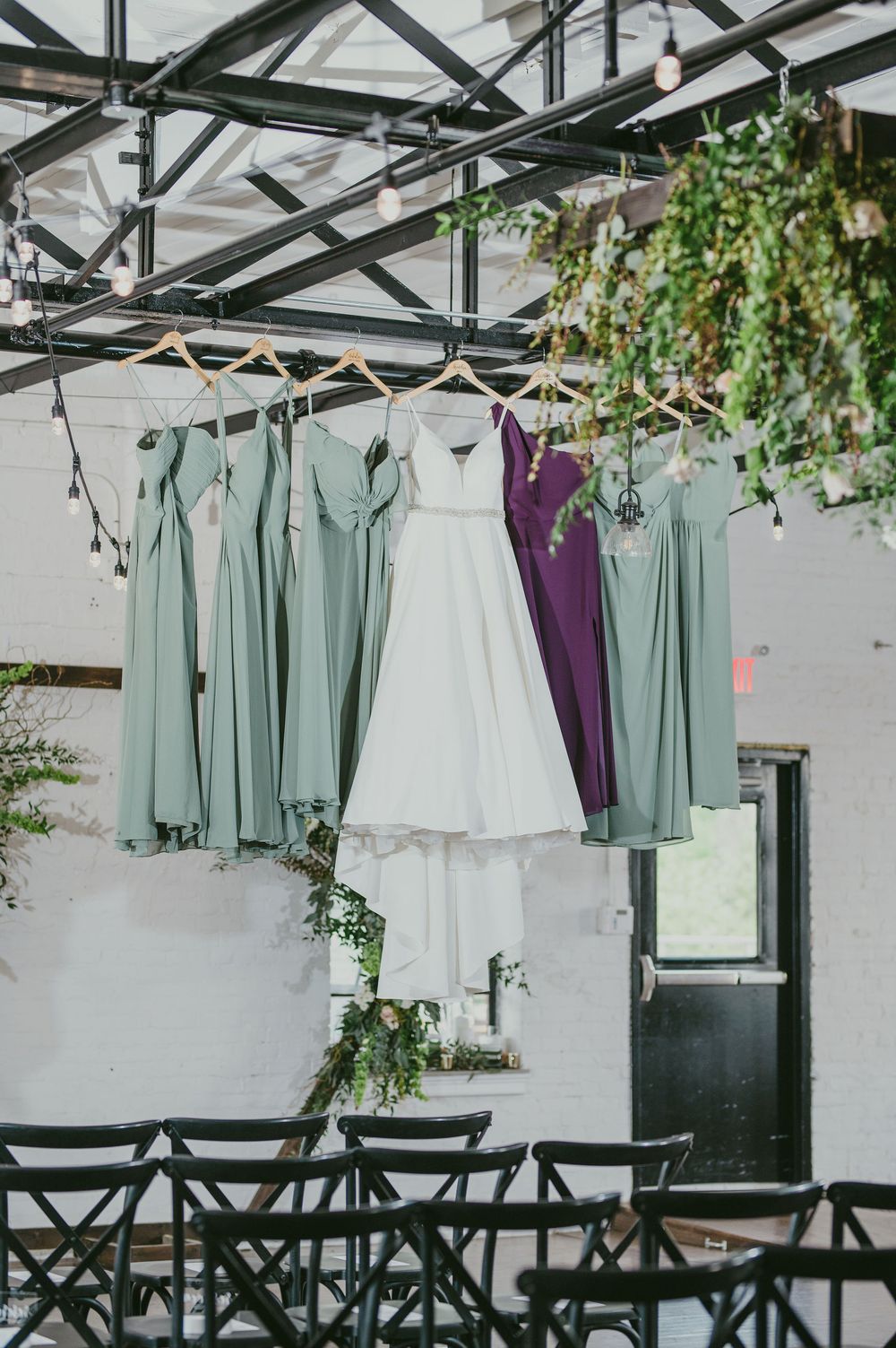 Elegant bridesmaid dresses in sage green and purple hang from a rustic ceiling with greenery in a wedding venue.