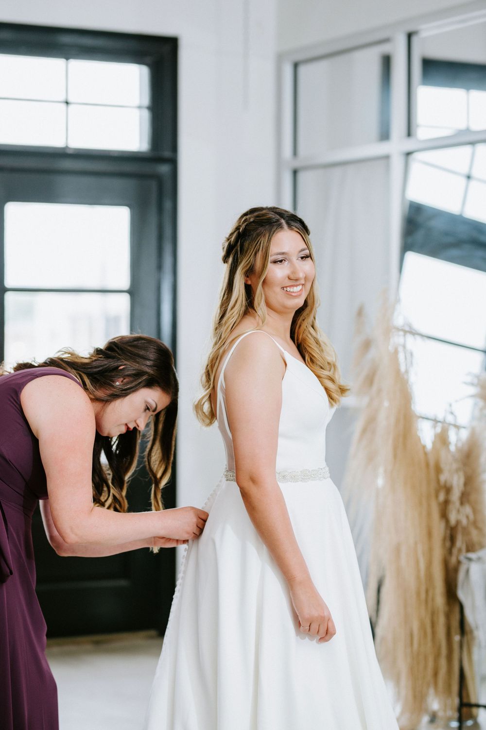 A group of women in purple bridesmaid dresses help zip up a white wedding gown in a bright sunlit room.