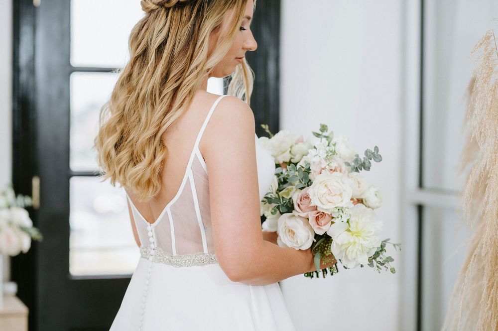 A bride in a white wedding dress holds a delicate bouquet of blush and white flowers against a bright window backdrop.