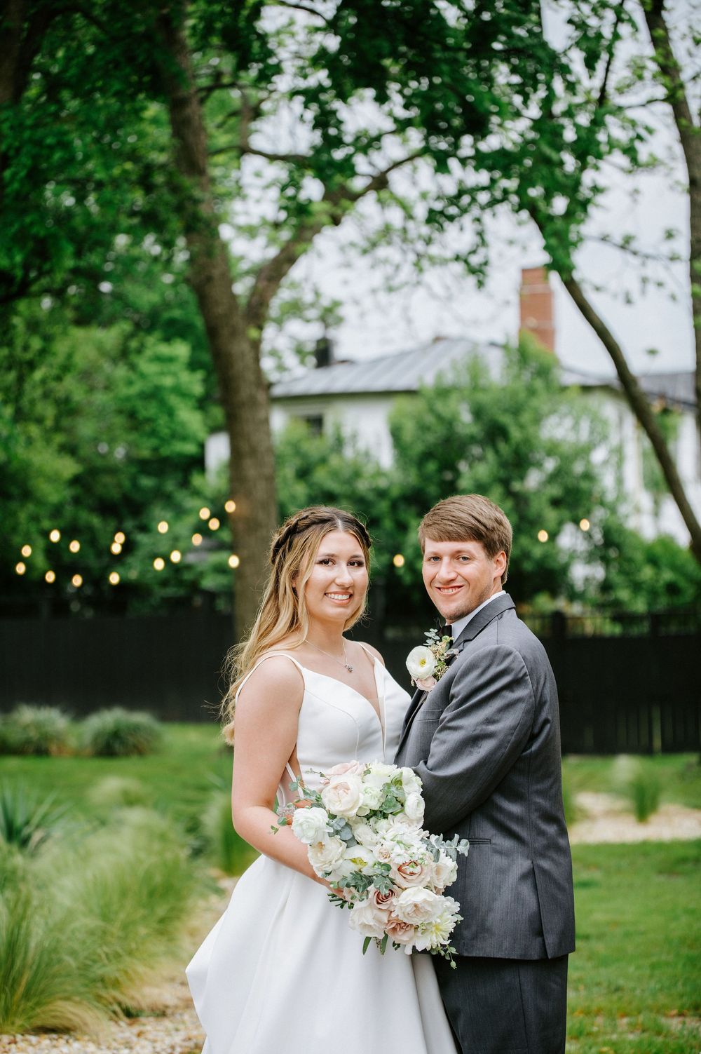 A couple in wedding attire poses together in a garden setting with string lights and greenery in the background.