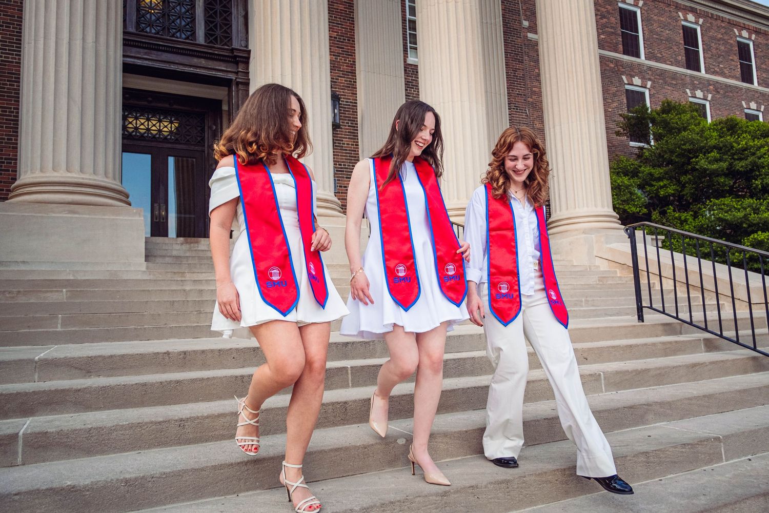 Three graduates in white dresses and red stoles pose on university building steps.