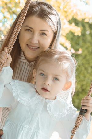 A smiling woman and a young blonde girl in white dress on a rope swing outdoors in autumn.