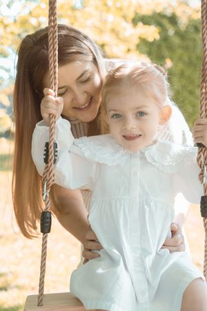 A smiling mother hugs her young blonde daughter sitting on a wooden rope swing outdoors in autumn.