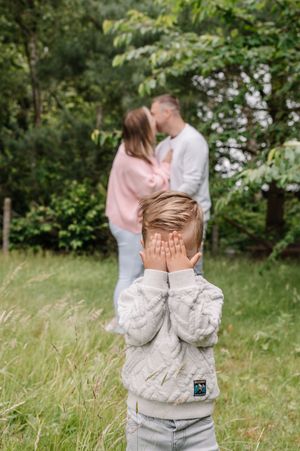 A young boy covers his eyes as his parents share a kiss in a lush green park behind him.
