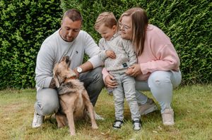 Family with toddler and German Shepherd dog posing together on green grass in a garden.
