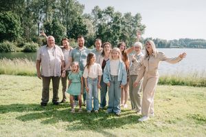 A joyful multigenerational family poses outdoors near a lake on a sunny day, smiling and waving together.