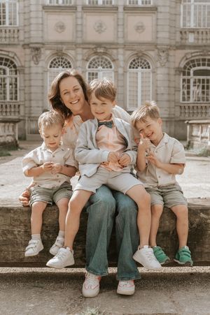 A smiling mother sits outdoors with her three young sons, all dressed in smart casual outfits, posing before a grand building.