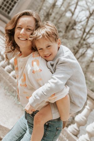 A smiling woman carries a happy young boy on her back outdoors on a sunny day.