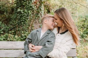 A mother and young son share a tender moment on a wooden bench surrounded by lush green foliage in autumn.