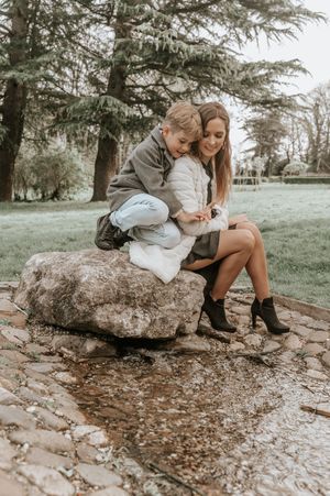 A woman and young child sit together on a large rock beside a stream in a lush green park.
