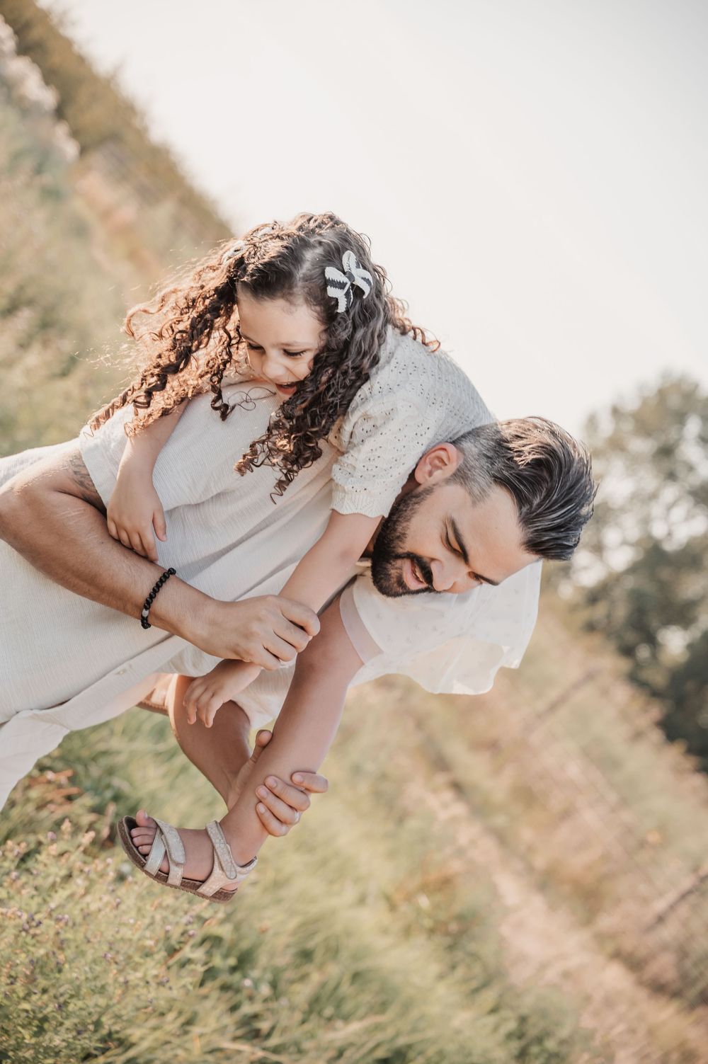 A bearded man gives a curly-haired girl a piggyback ride in a sunny field, both laughing joyfully.