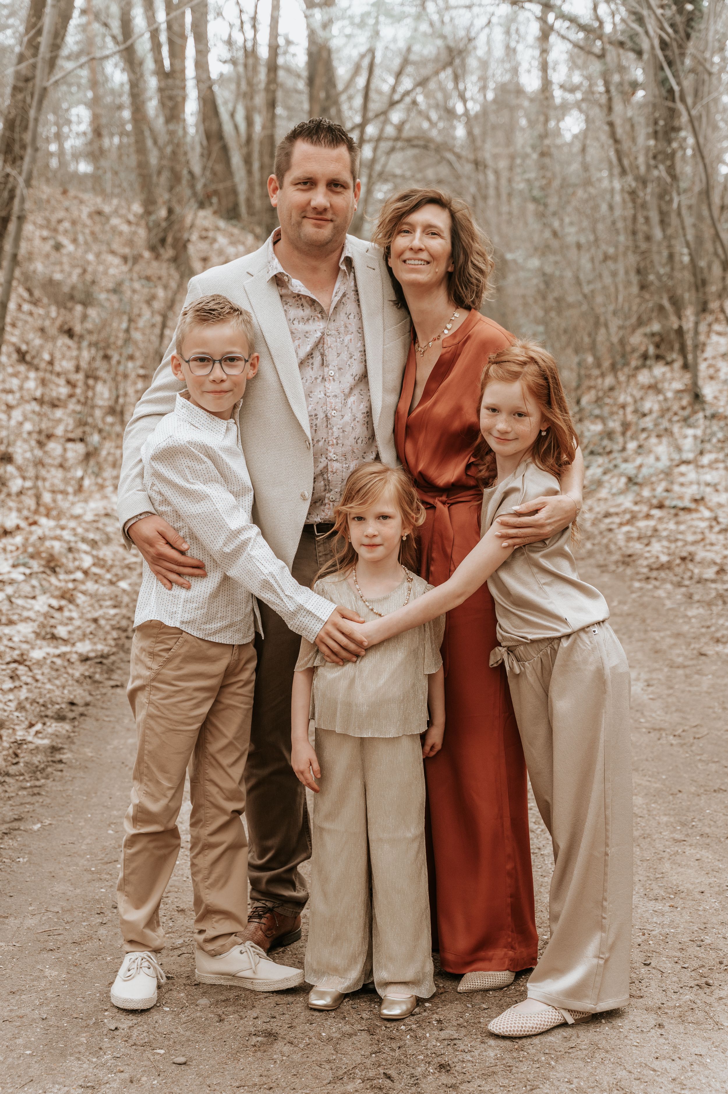 Family of five posing together on a wooded path in coordinated neutral and rust-toned outfits for a professional portrait.
