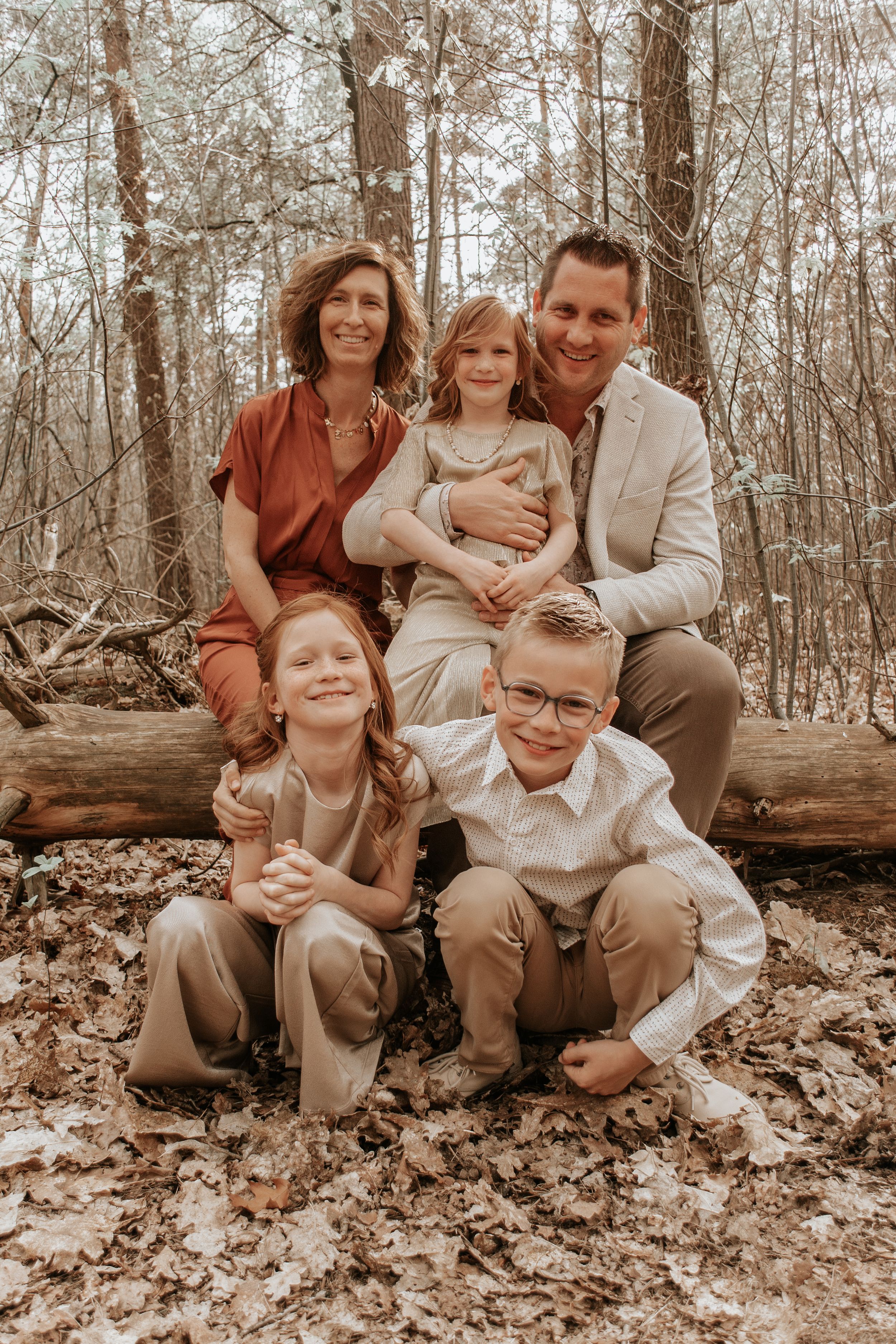 Family of five posing in autumn woods, parents seated on log with three children, wearing neutral earth tones.
