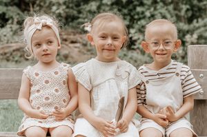 Three young children smiling and sitting together on a wooden bench outdoors in summer clothing.