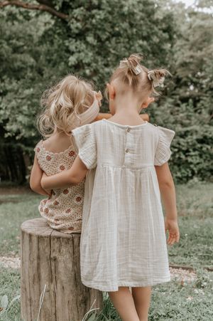 Two toddler girls in summer dresses stand with backs turned, looking at lush green trees outdoors.