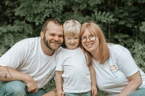 Happy family of three in matching white t-shirts smiling outdoors with lush green foliage background.