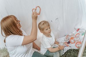 A mother and young boy craft together in a white teepee, painting colorful rings on fabric outdoors.