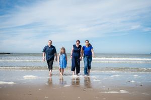 Family of four walking barefoot on a sandy beach with ocean waves, wearing blue outfits on a sunny day.