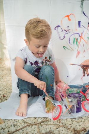 Young boy painting with colorful paints, sitting on white sheet with paintbrush, wearing paint-splattered white shirt.