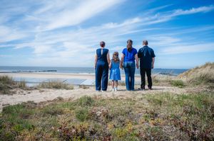 Family of four standing on sandy beach dunes overlooking the ocean on a sunny day with blue skies.