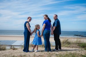 Family of four in blue outfits holding hands on a sandy beach with ocean and blue sky backdrop.