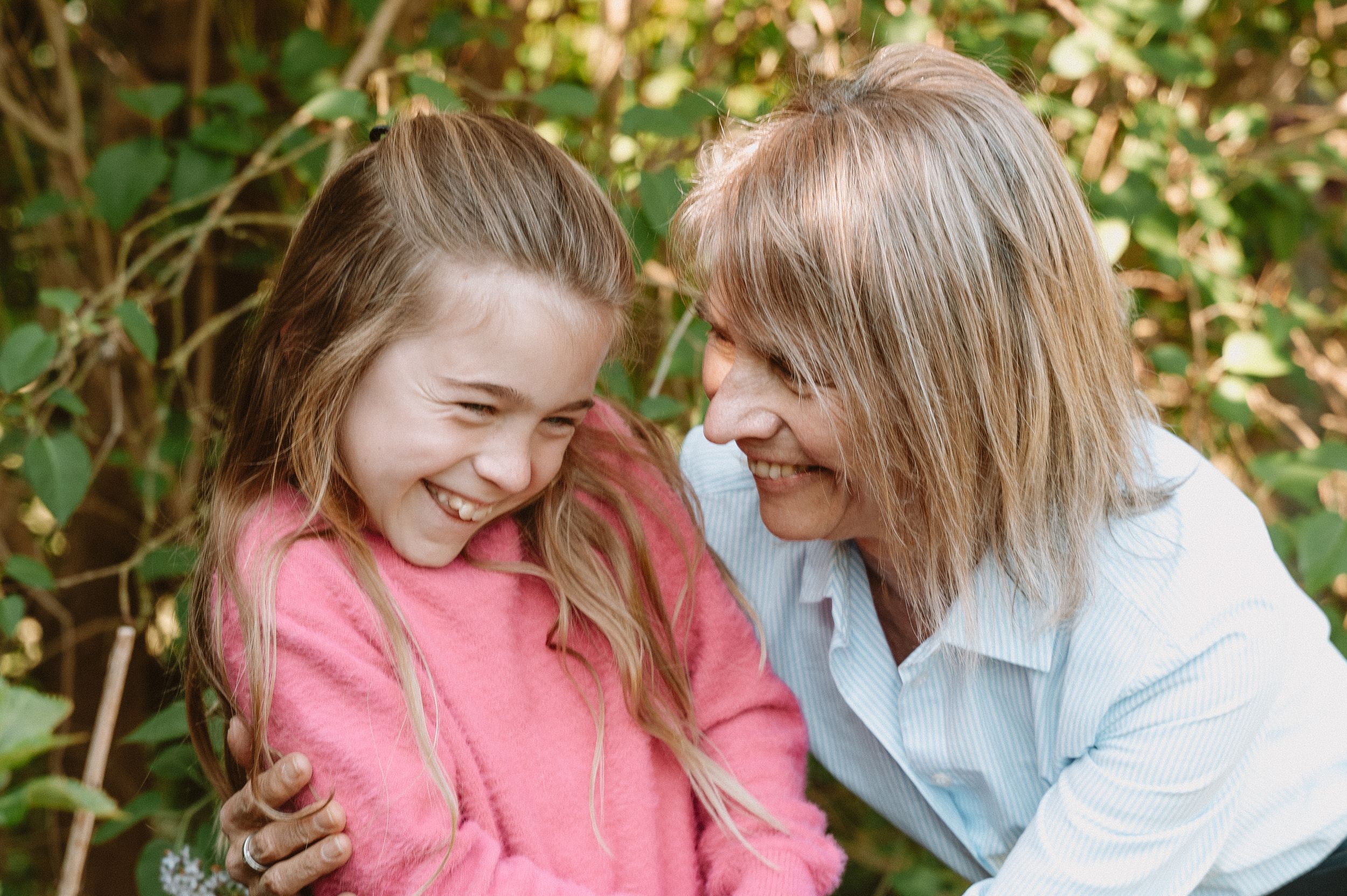 A woman and young girl laughing together outdoors, sharing a joyful moment in front of green foliage.