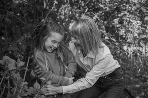 Two girls laughing together outdoors among lush foliage in a candid black and white portrait photo.