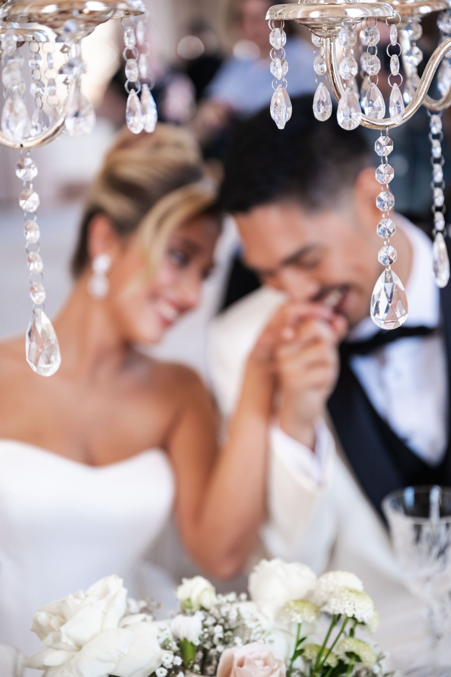 Bride and groom hold hands beneath sparkling crystal chandelier at elegant wedding reception table.