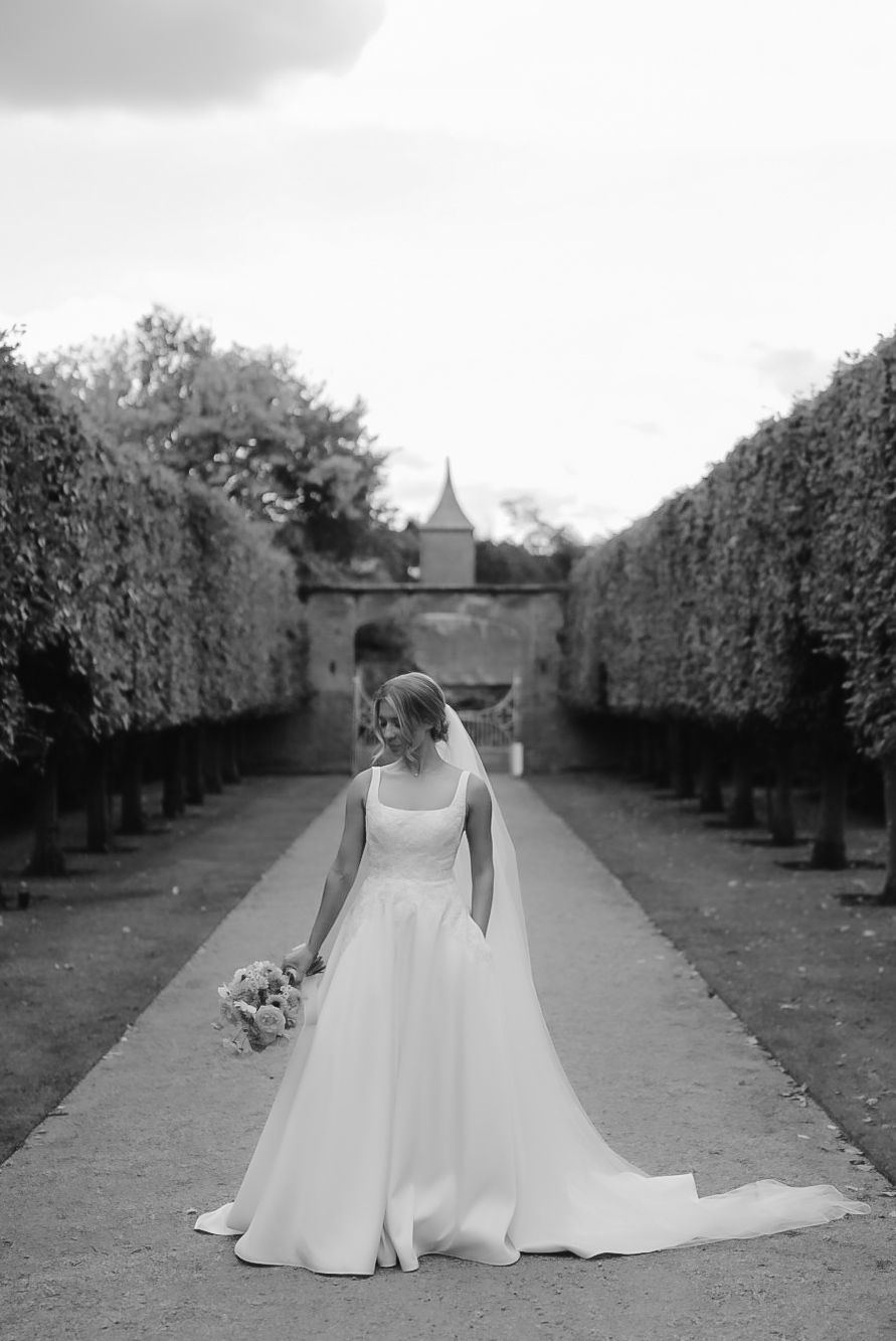 A bride in a flowing white gown poses gracefully in a garden pathway lined with manicured hedges.
