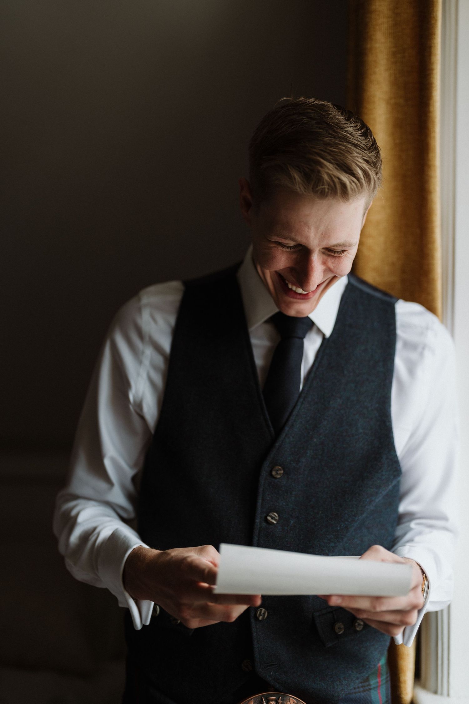 A person in formal attire reads a document by a window with warm lighting.