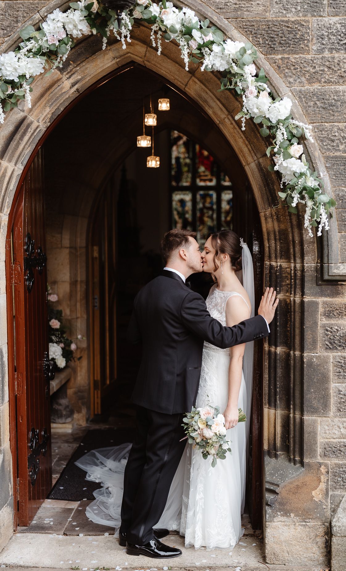 A romantic moment unfolds under a stone archway decorated with white floral arrangements.