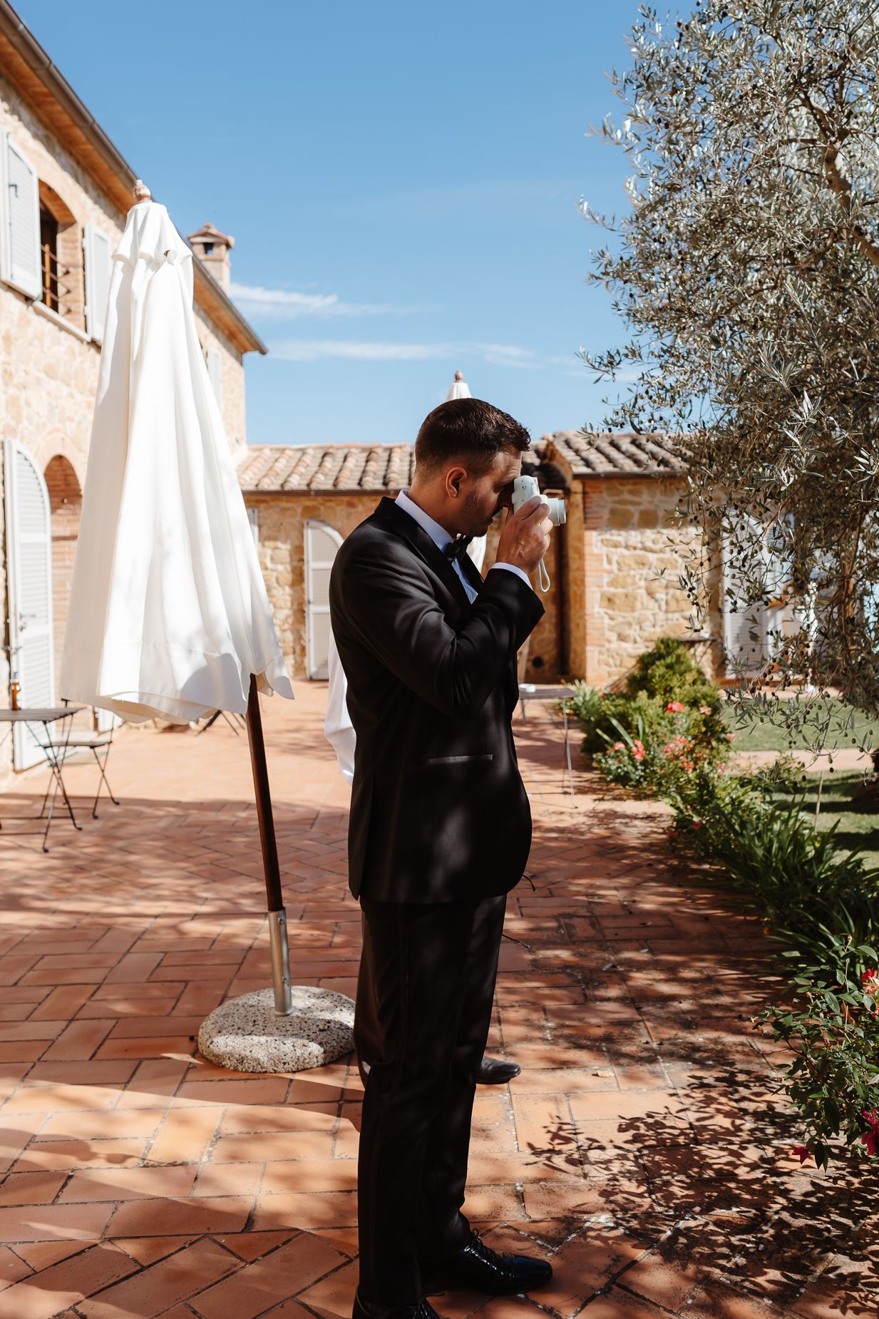 A person in formal attire takes photos on a Mediterranean-style terrace with terra cotta tiles.