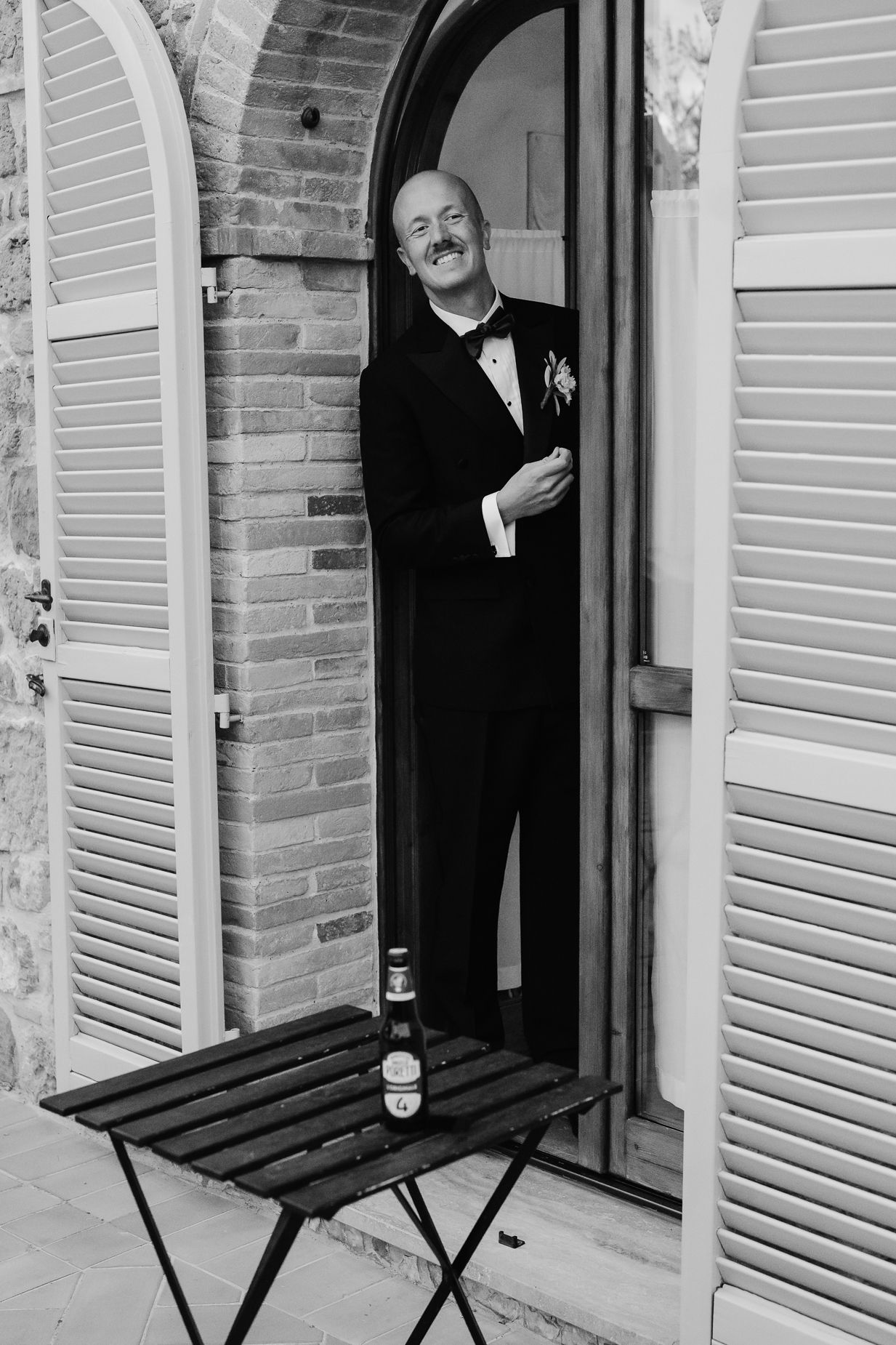 A person in formal attire stands in a doorway of a Mediterranean building with shuttered windows.