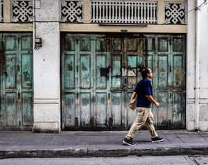Person in a blue shirt and khaki pants walking past a rustic teal door on a city street.
