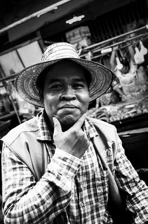 Man in straw hat resting chin on hand at busy market stall in black and white portrait.