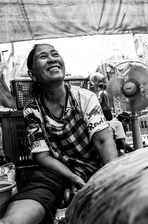 Smiling Southeast Asian woman in checkered shirt sitting near electric fan at market.