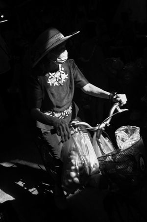 Woman in hat and face mask riding bicycle with basket in dramatic black and white light.