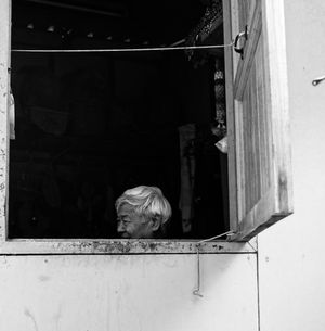 Elderly person with white hair peering out of weathered wooden window in black and white.