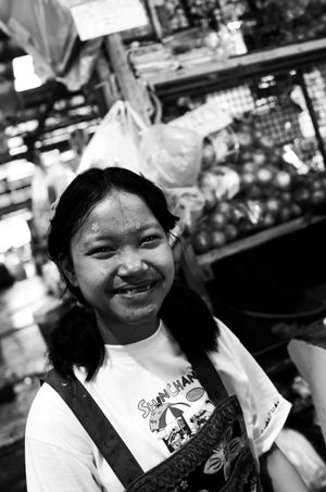 Young smiling girl in white shirt at busy outdoor market in black and white photo.