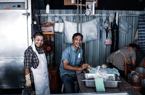 Two smiling workers in aprons inside a small kitchen food stall.