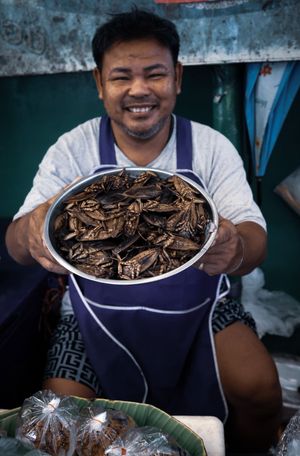 Smiling vendor holding large bowl of roasted insects at outdoor market stall.