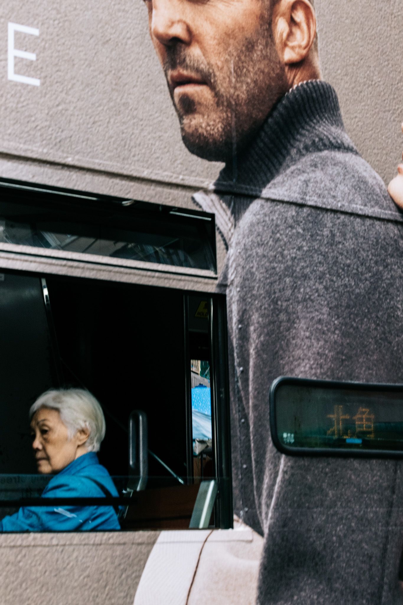 A man in a grey turtleneck stands beside a bus with an elderly woman visible through the window.