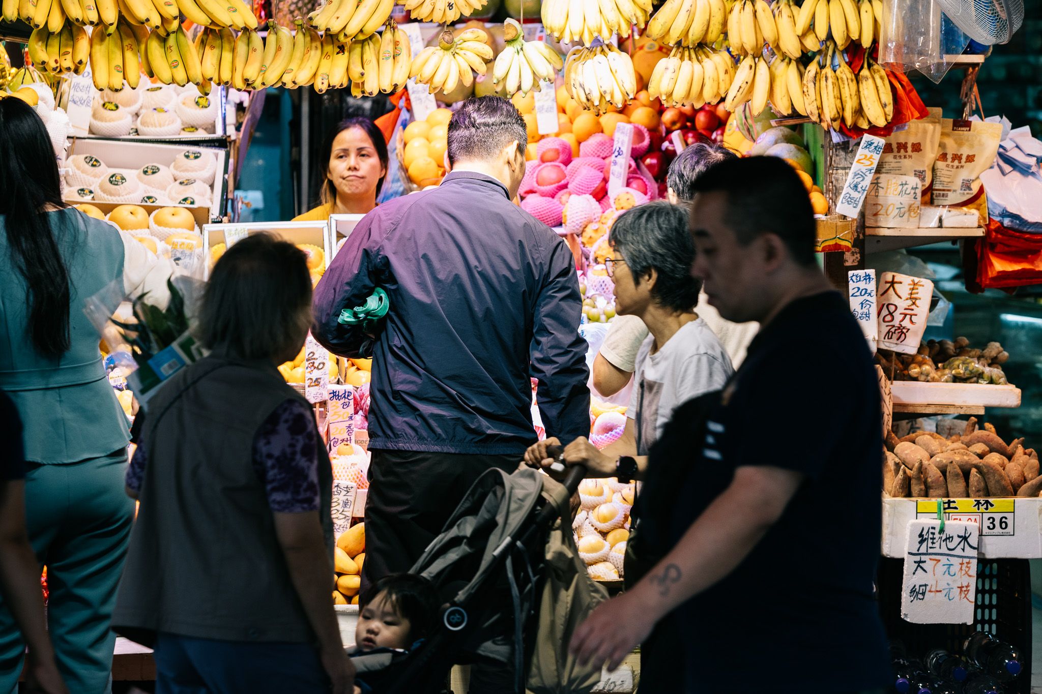Crowds of people browse a vibrant outdoor fruit market stall with hanging bananas in Hong Kong.