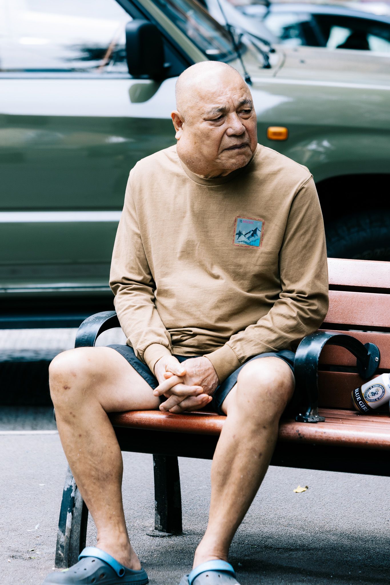An elderly bald Asian man sits alone on a bench with hands clasped, deep in thought outdoors.